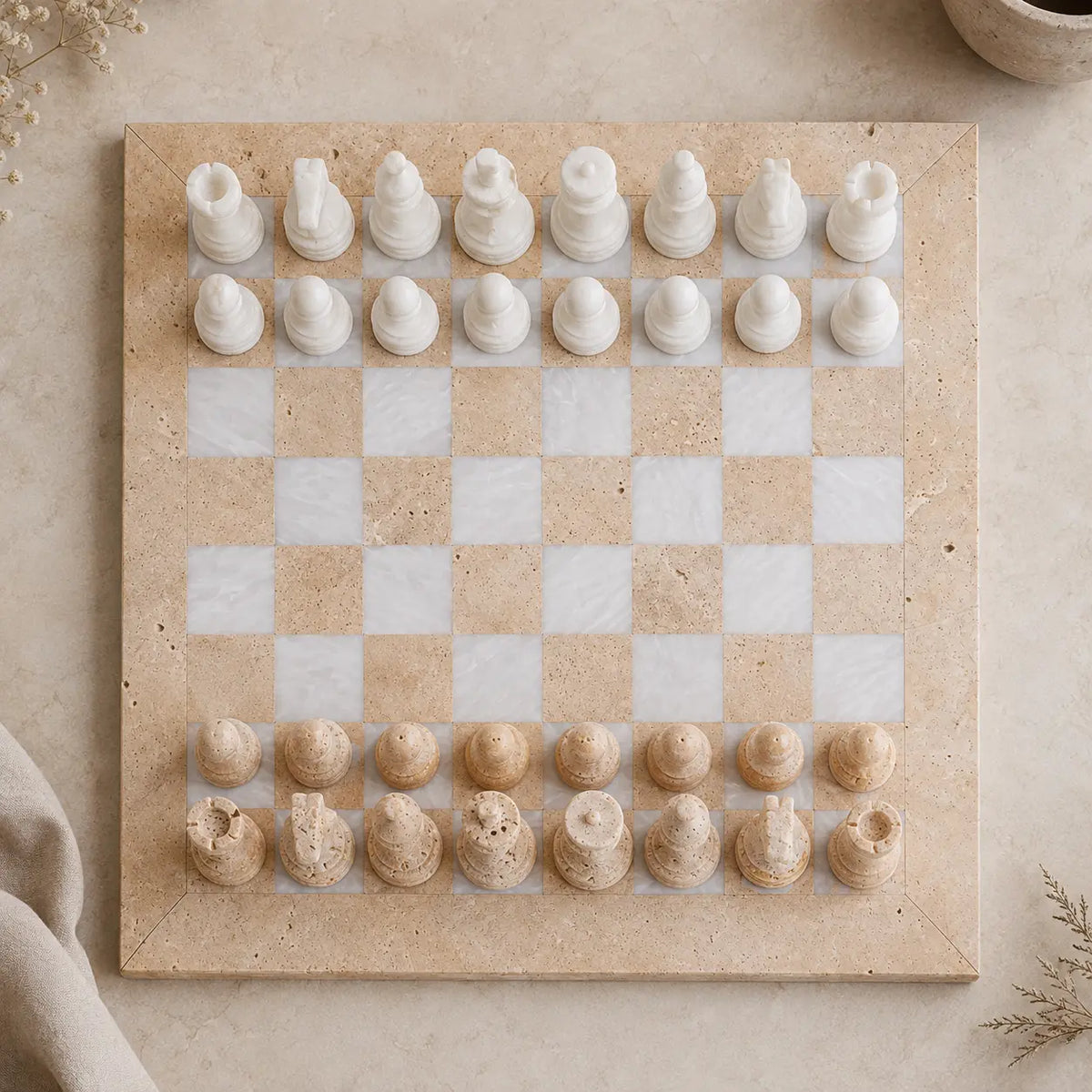 A square stone chessboard with white and beige checkered squares, topped with white and beige chess pieces, arranged on a neutral stone surface.