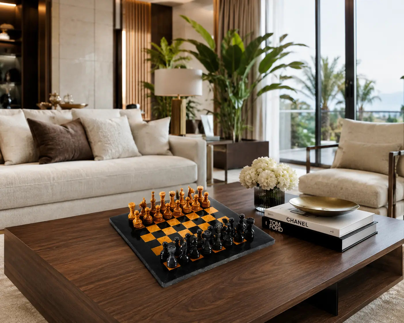 A modern living room interior with a dark wood coffee table in the foreground holding a black-and-gold chessboard and chess pieces, books, and flowers. A cream sofa, armchair, plants, and large windows are visible in the background.