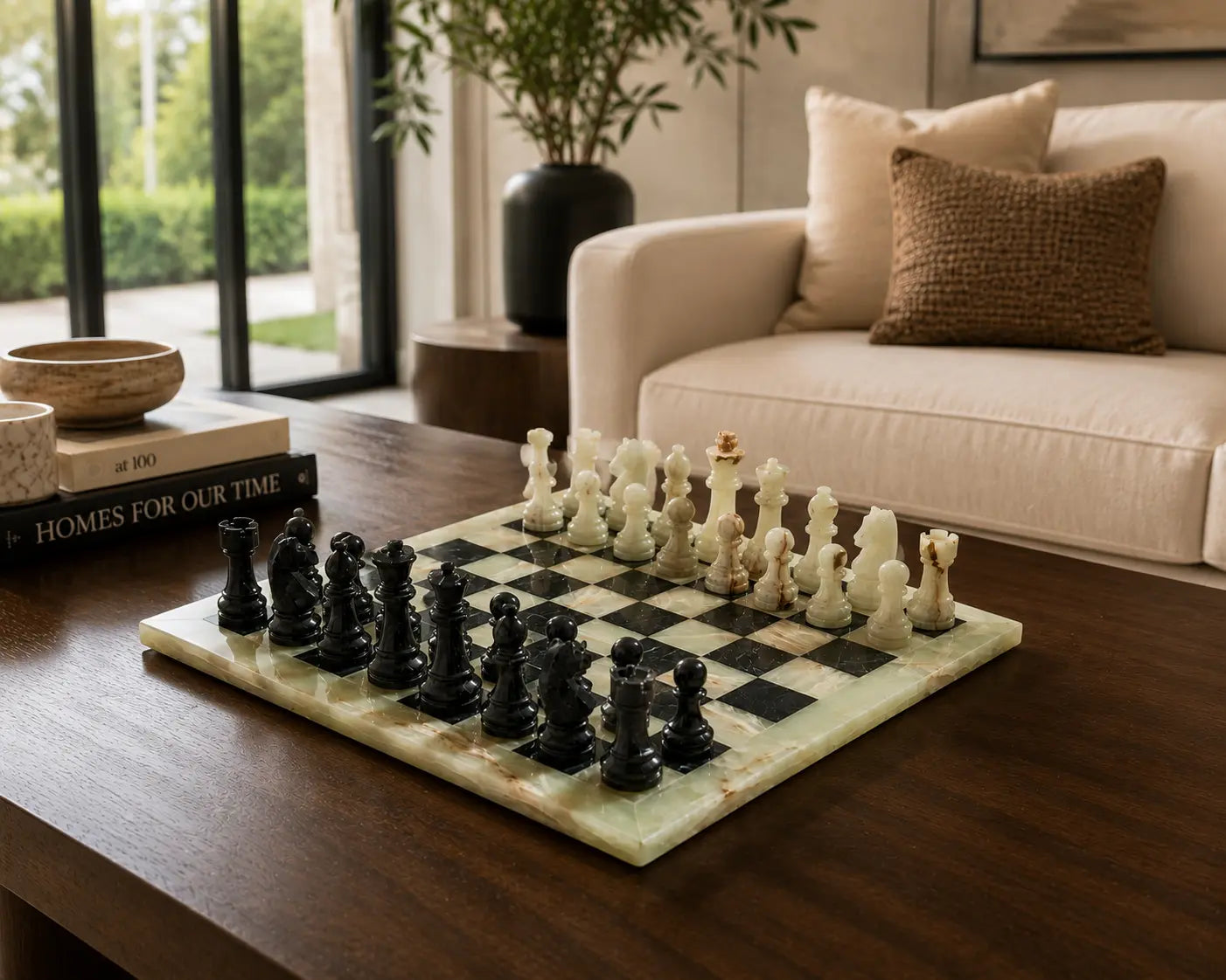 A marble chessboard with black and cream chess pieces set on a dark wooden table in a modern living room with a beige sofa and large windows.