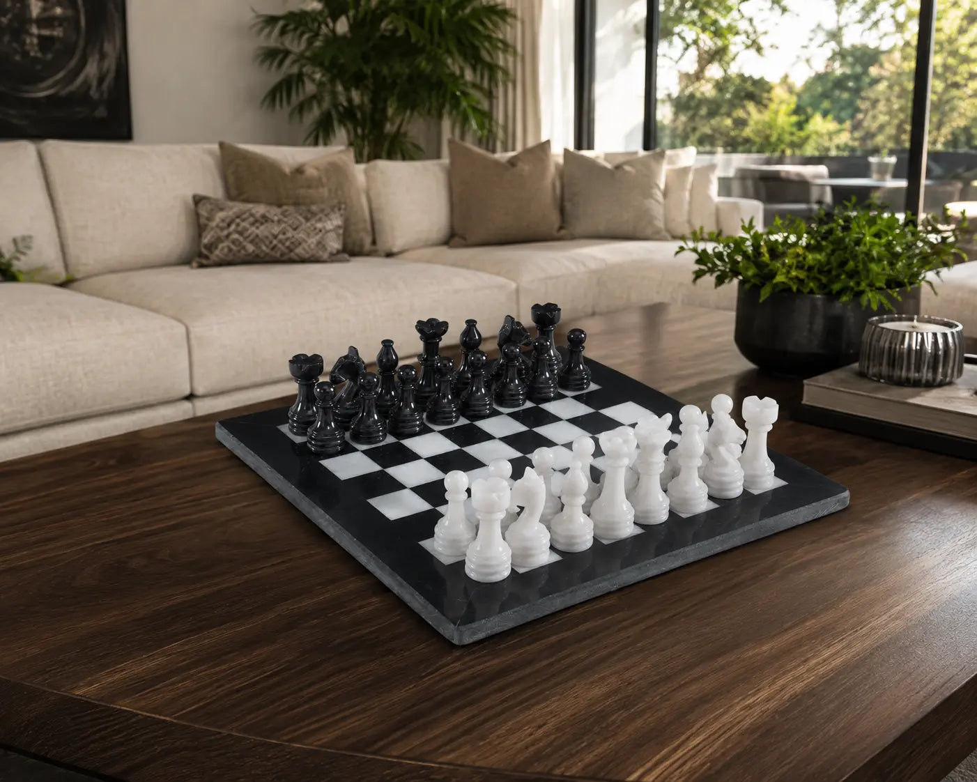 A black and white checkers game set on a dark gray slate board, styled on a dark wood coffee table in a modern living room with a beige sofa and plants.