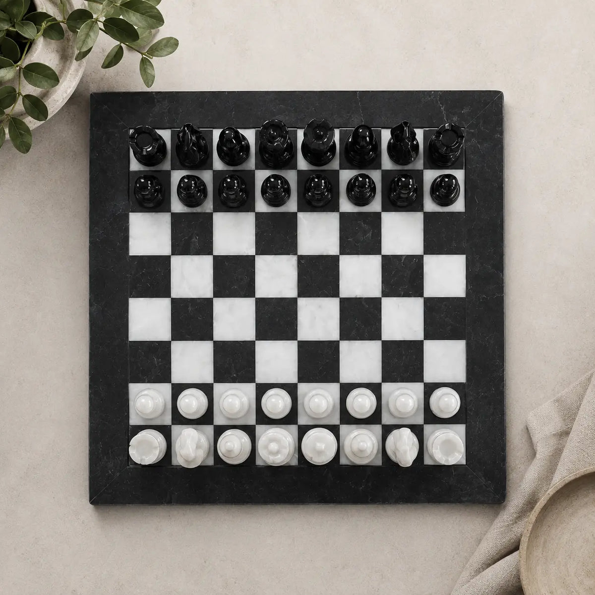 A black and white marble chessboard with carved black and white chess pieces, photographed from above on a light beige surface with small decorative plant leaves and fabric props at the edges.