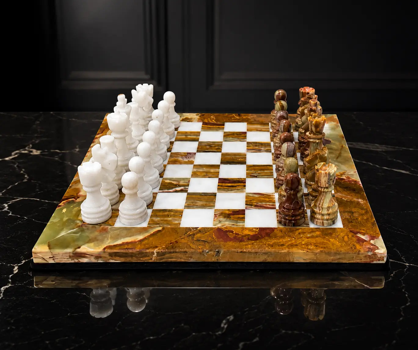 A luxury marble chess set on a polished black marble surface against a dark paneled background. The chessboard features alternating white and brown/bronze checkered squares, with white chess pieces on one side and intricately carved brown, gold, and green stone chess pieces on the other.