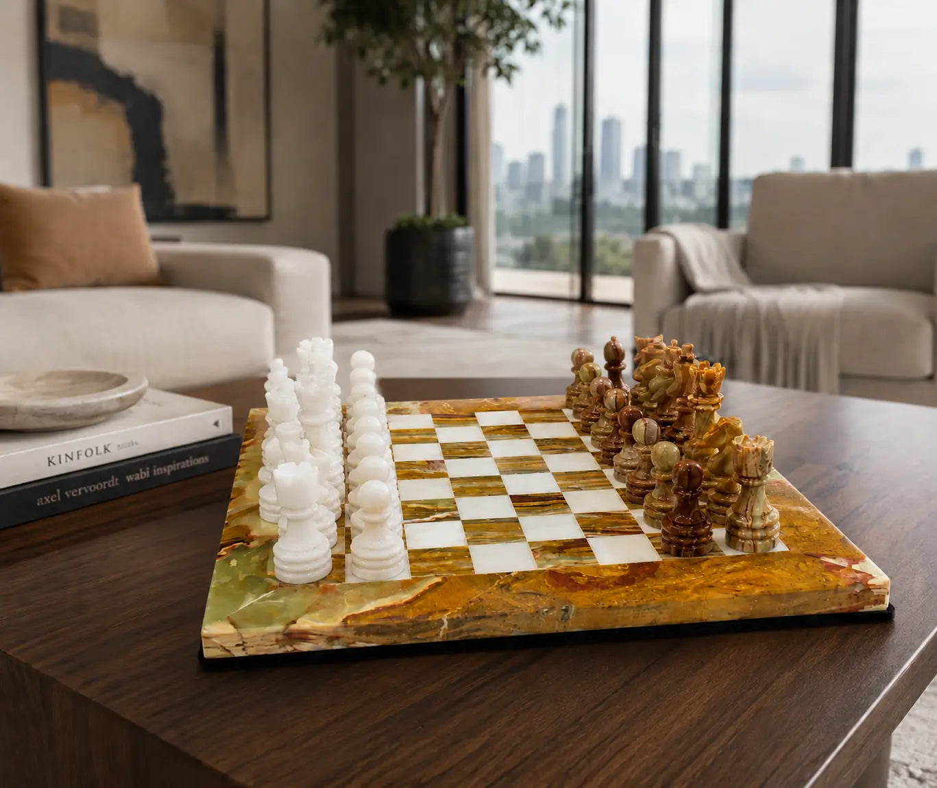 A luxury marble chessboard set on a dark wood table in a modern living room. The chessboard features a polished gold and green marble base with white and brown carved wooden chess pieces arranged on it.