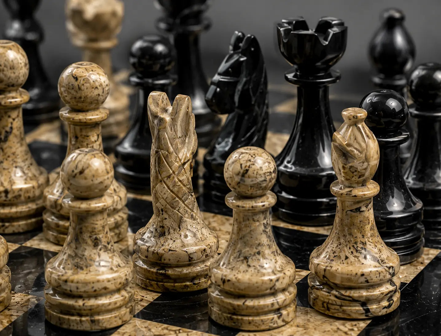 Close-up of a polished stone chess set on a dark and light marble chessboard, featuring cream-colored pieces with black marbling and glossy black pieces in the background.