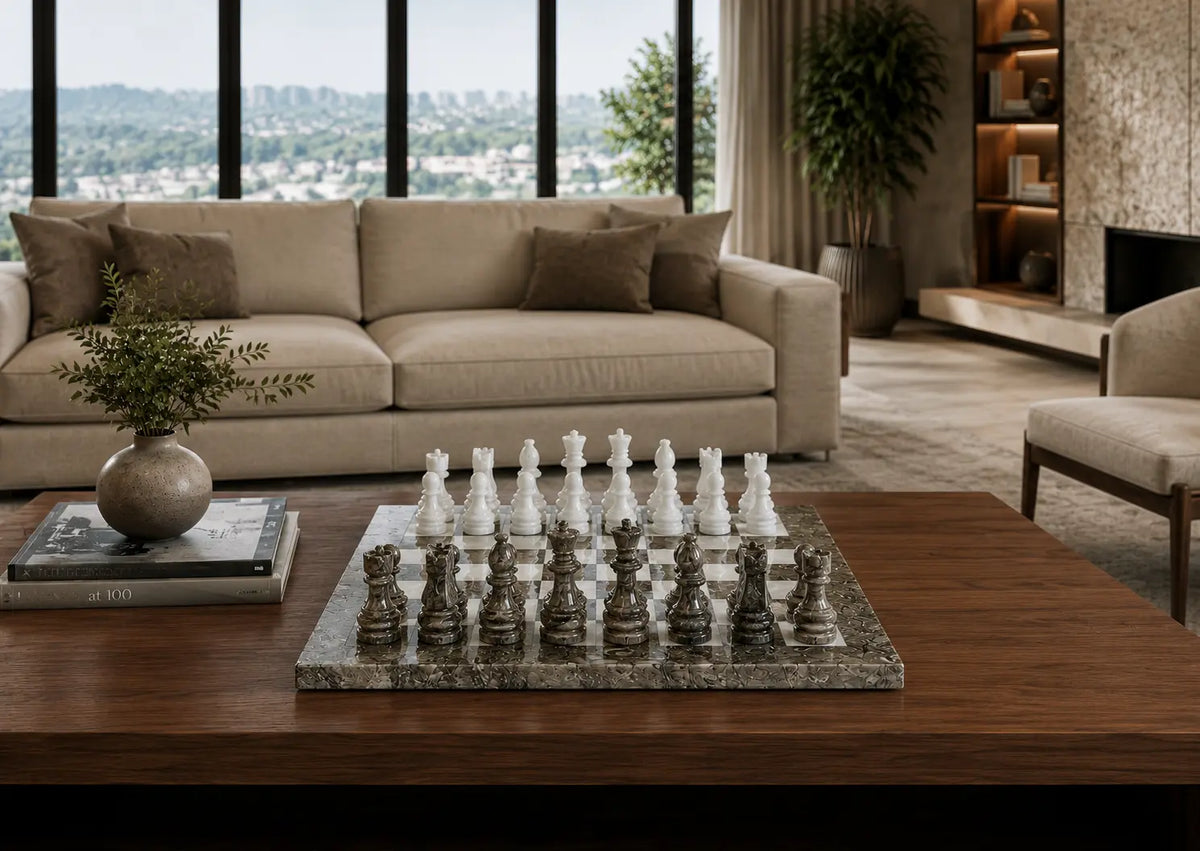 A marble chessboard with white and gray chess pieces set on a dark wood coffee table in a modern living room with a beige sofa and large windows.