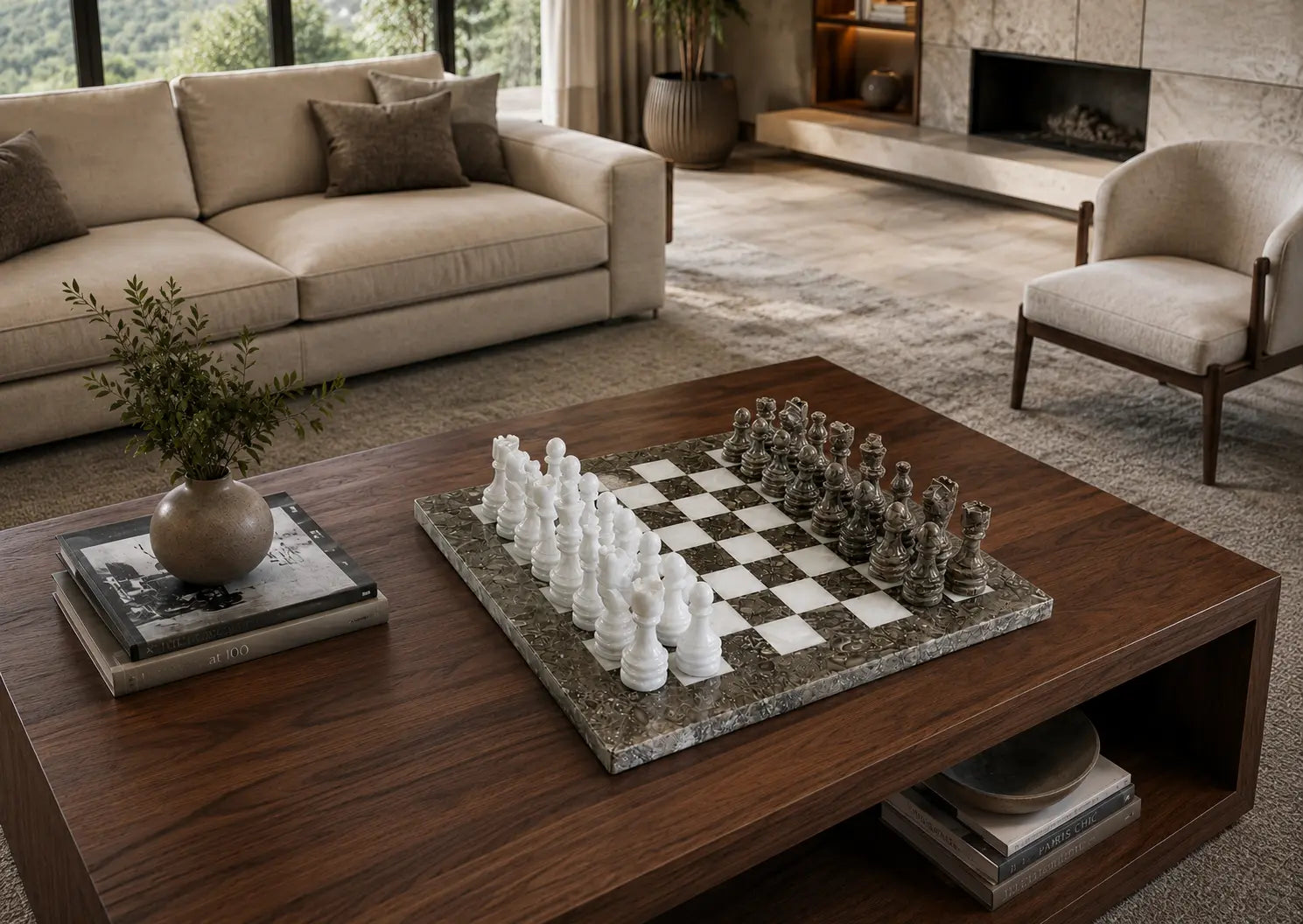 A dark wood coffee table in a modern living room setting, featuring a marble chessboard with white and dark chess pieces. The table is styled with books and a small potted plant, with a beige sofa and a fireplace visible in the background.