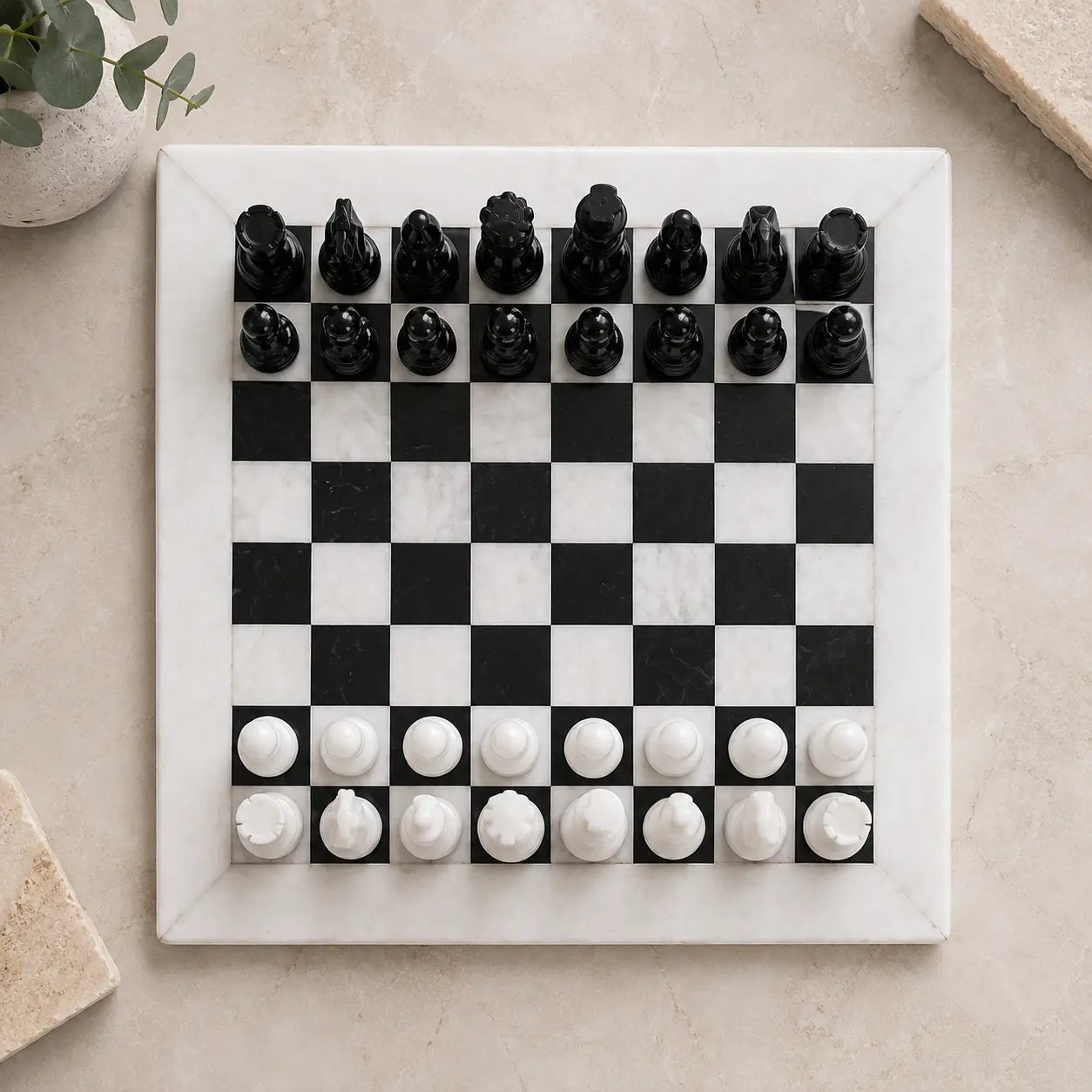 A square black-and-white checkered chessboard made of polished stone, with black and white chess pieces arranged at the bottom, photographed from above on a light beige surface with small decorative props in the corners.