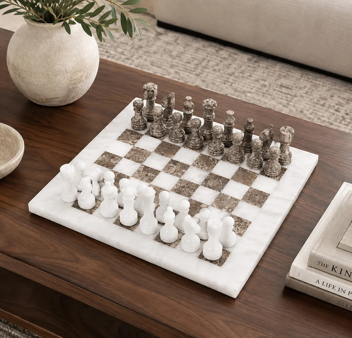 A marble chess set with white and gray stone chess pieces arranged on a white and gray checkered marble board, placed on a dark wood coffee table in a styled living room setting.