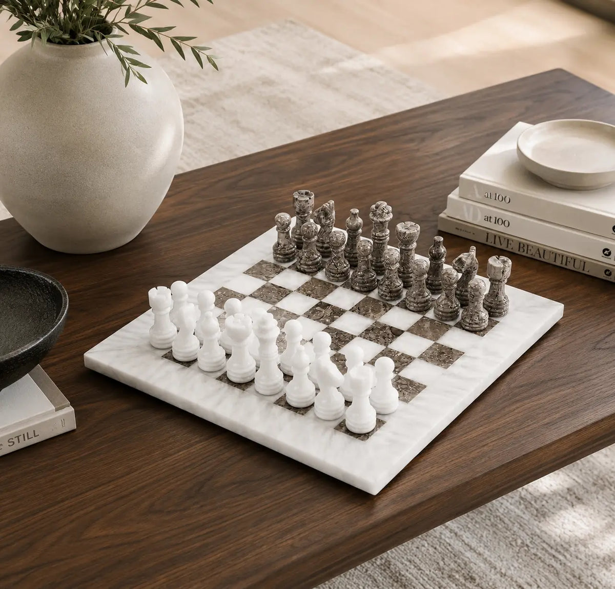 A marble chess set with white and dark gray pieces on a white-and-dark marble board, styled on a wooden table with a vase, books, and a bowl nearby.